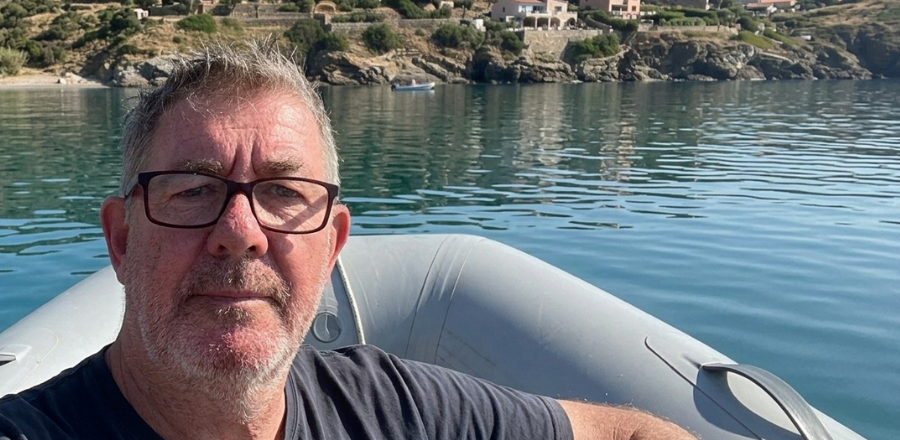 Greg Berryman sits in a grey inflatable dinghy, positioned to the left of frame, with a calm bay, rocky coastline, and hillside village in the background.