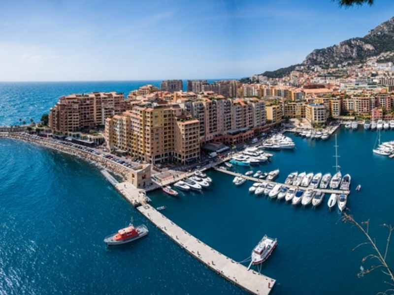 Wide aerial view of Monaco's Fontvieille harbour, showing the curved breakwater, small boats near the pier, and waterfront apartment buildings backed by mountains.