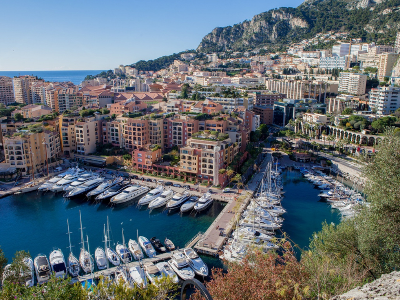 Aerial view of Fontvieille marina in Monaco, with rows of yachts docked along the harbour and dense hillside apartment buildings rising behind.